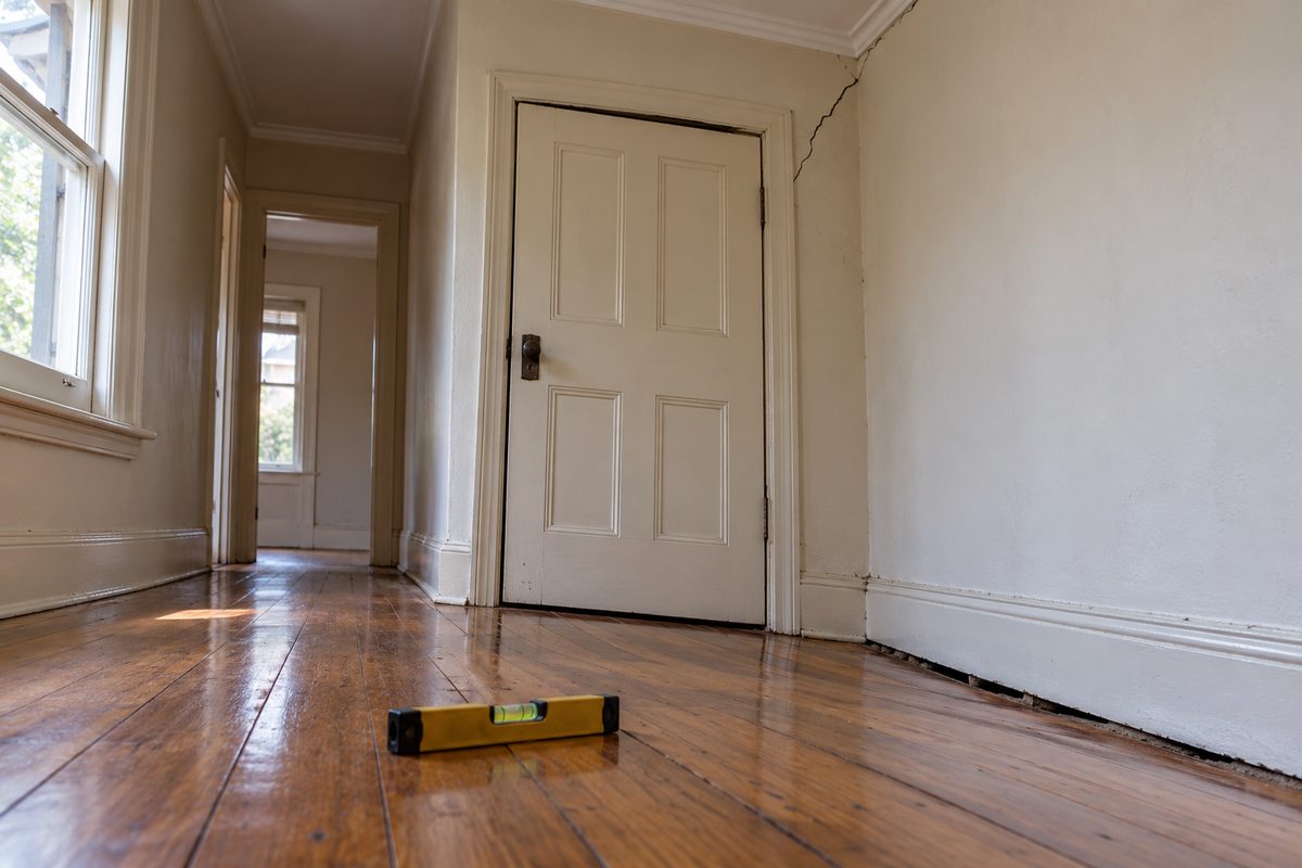 Hallway of an older Albury weatherboard home, floorboards dipping toward one wall, a door out of square with an uneven gap at the top, a diagonal plaster crack from door frame to cornice, a gap under the skirting, a spirit level with bubble off centre.